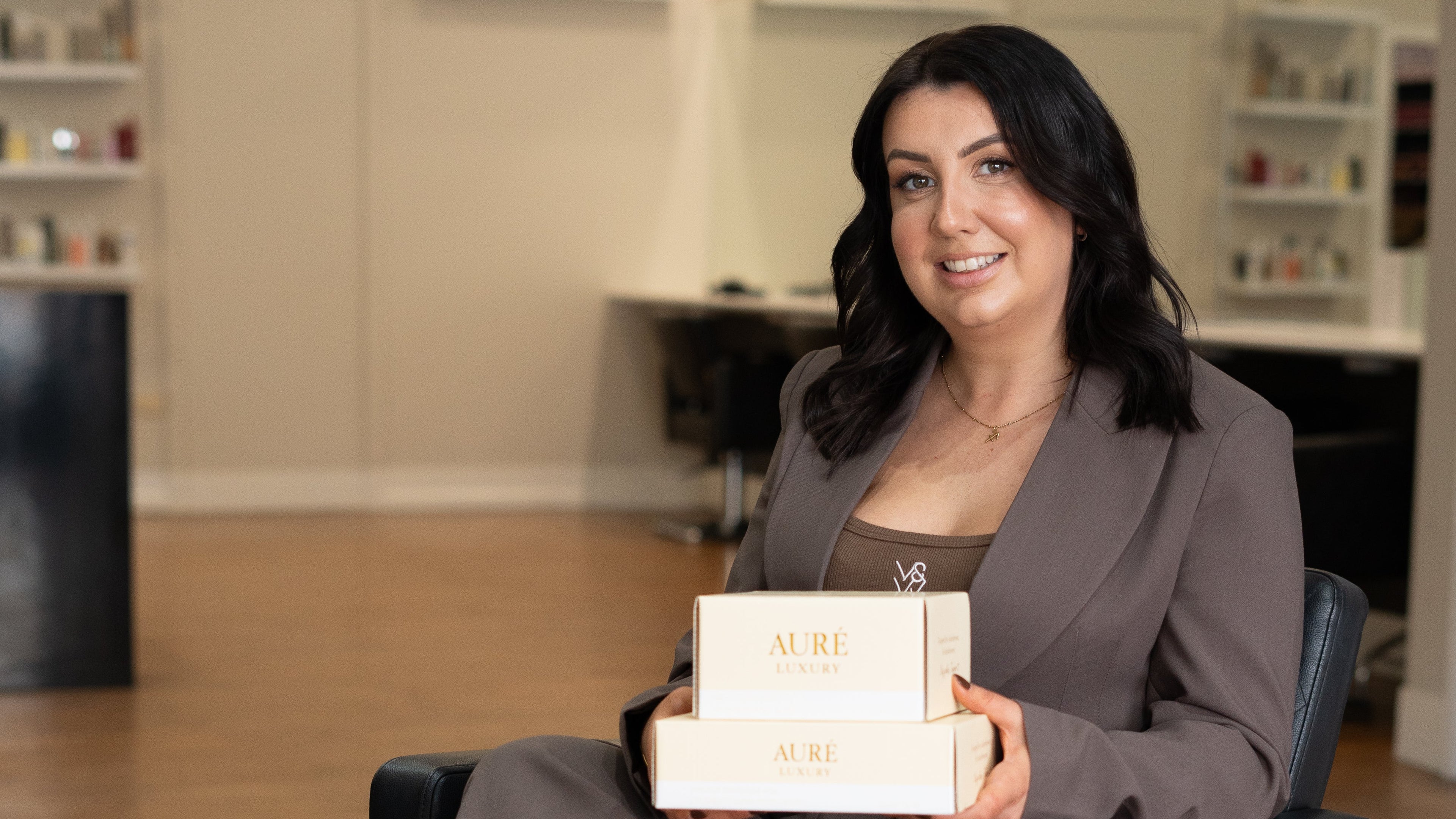 Woman sitting in a salon chair holding a box, with a blurred salon interior in the background.